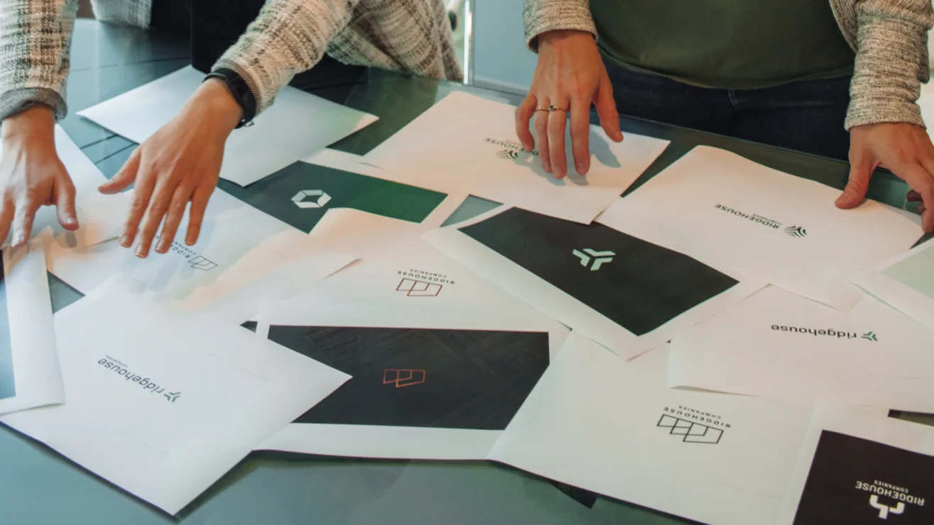 People reviewing printed logo concept proofs scattered on a glass conference table.