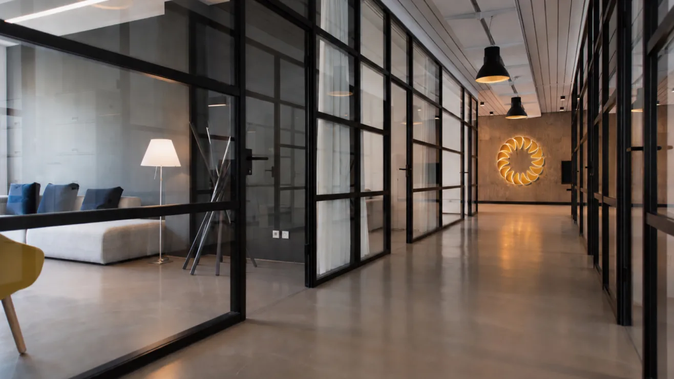A modern office hallway featuring glass partition walls and a glowing yellow circular logo mounted on the end wall.