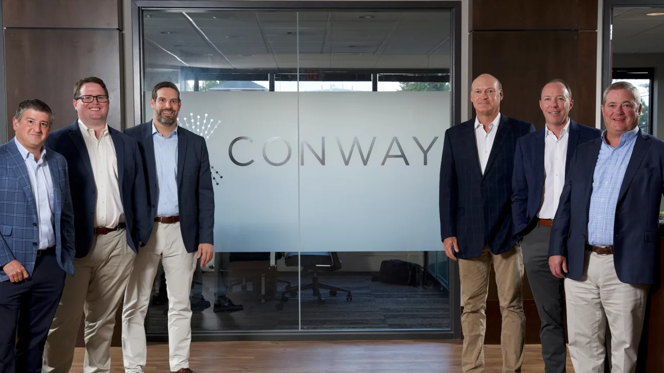 Six team members in business attire posing in front of a frosted glass office wall featuring the Conway logo.