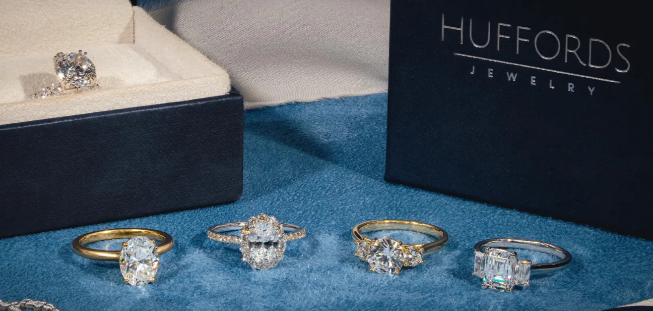 A row of four diamond rings displayed on a plush blue surface in front of a Huffords Jewelry box. A singular ring displayed inside a navy blue box.